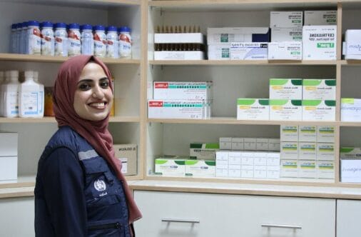 A woman in a WHO vest smiles in front of medical supplies.